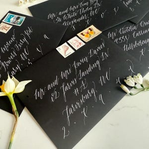 May include: Black envelopes with elegant white calligraphy, addressed with names and addresses. Decorative stamps are affixed to some envelopes. A white daffodil and small white flowers are arranged around the envelopes.