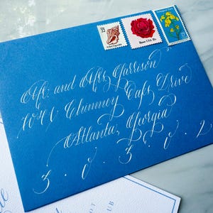 May include: A blue envelope with white calligraphy and three postage stamps. The stamps feature a seashell, a red rose, and yellow flowers. The calligraphy includes a name, address, and city.