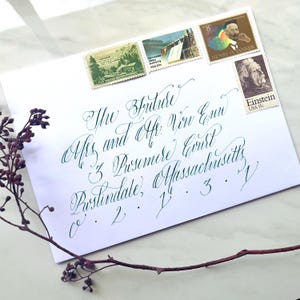 Könnte beinhalten: Ein weißer Umschlag mit grüner Kalligraphie, adressiert an "The Future" mit Briefmarken, die einen Staudamm, Henry O. Tanner und Einstein zeigen. Ein Zweig getrockneter Blumen liegt auf der Oberfläche.