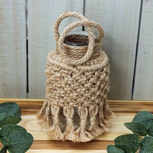May include: A glass vase wrapped in natural jute twine with a knotted handle and fringe detail. The vase is sitting on a wooden surface with green foliage.