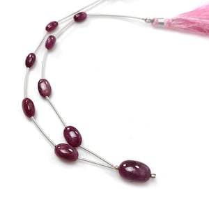 Könnte beinhalten: Eine Halskette mit ovalen, tiefroten Edelsteinen, die auf dünnem, silberfarbenem Draht aufgereiht sind. Die Halskette ist mit rosa Quasten versehen. Die Edelsteine haben ein poliertes, glänzendes Aussehen, vor weißem Hintergrund.