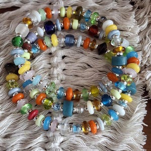 Peut inclure: Plusieurs bracelets de perles colorées. Les bracelets présentent des perles de verre dans des tons orange, jaune, vert, bleu et transparent, enfilées avec des entretoises argentées. Les bracelets sont présentés sur une surface texturée blanc cassé.