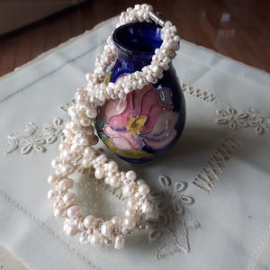 May include: A white pearl necklace draped around a dark blue vase with a floral design. The necklace is composed of numerous small, irregular pearls. The vase sits on a light-colored, embroidered cloth.