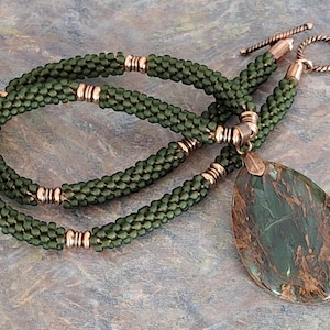 May include: A handcrafted necklace featuring a green beaded rope chain with gold-colored accents. The pendant is a large, teardrop-shaped stone with green and brown marbling. The necklace has a copper-colored clasp.