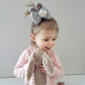 K&ouml;nnte beinhalten: Ein kleines Kind in einem rosa Pullover h&auml;lt ein braunes Pl&uuml;schkaninchen mit rosa-wei&szlig;en gestreiften Beinen. Das Kind tr&auml;gt ein graues gestricktes Hasenohrenband mit einem wei&szlig;en Bommel.