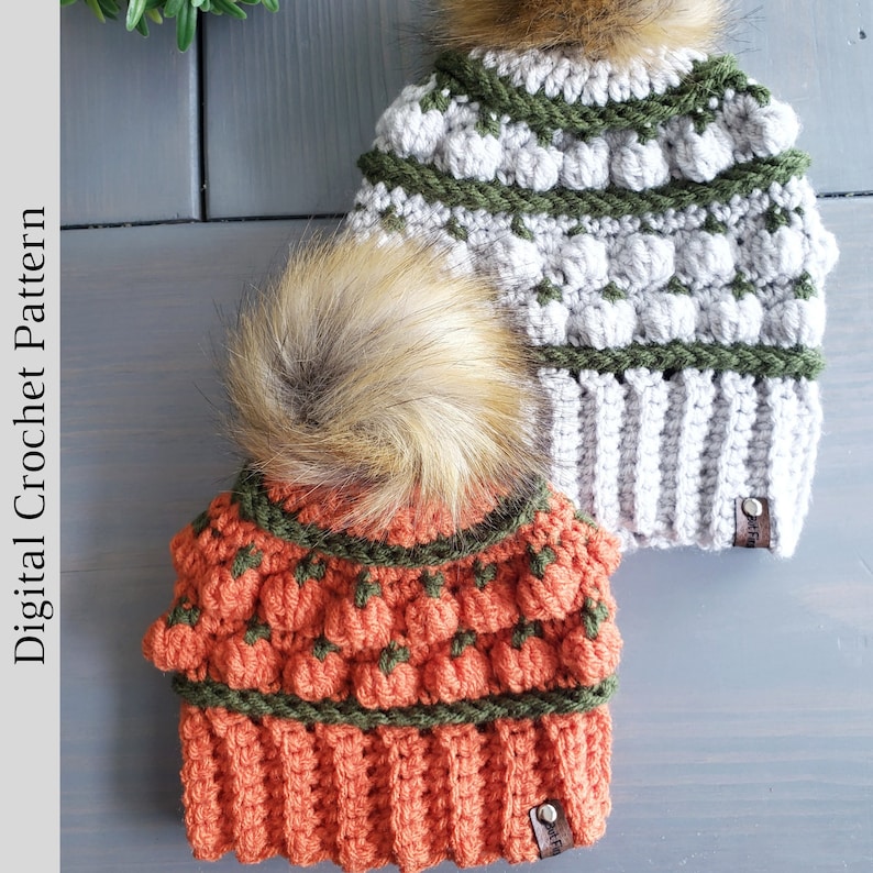 May include: Two crocheted hats with pom poms. One hat is orange with a green and orange pattern and the other is gray with a green and gray pattern. Both hats have a brown faux fur pom pom.