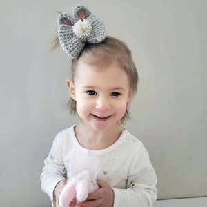 K&ouml;nnte beinhalten: Ein kleines Kind mit kurzen braunen Haaren tr&auml;gt ein wei&szlig;es Langarmshirt. Das Kind h&auml;lt ein rosa Stofftier-Kaninchen. Das Kind tr&auml;gt ein graues geh&auml;keltes Hasenohren-Haarband mit einem wei&szlig;en Bommel.