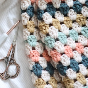 May include: A crocheted blanket with a granny square pattern in shades of beige, peach, teal, and light green.