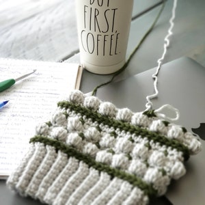 May include: A close-up of a crocheted hat with a green and white color pattern. The hat has a textured surface with raised bumps. The hat is lying on a table next to a white mug with the text "But First, Coffee" printed on it.