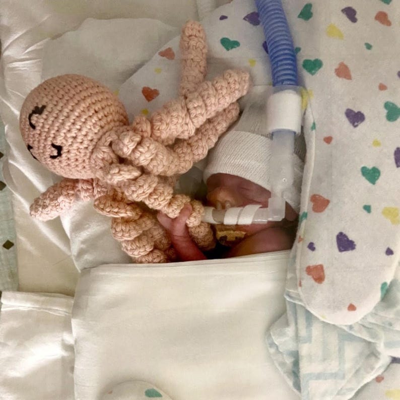 Puede incluir: Un juguete de pulpo rosa tejido junto a un beb&eacute; en una cama de hospital. El beb&eacute; lleva un gorro blanco y tiene tubos m&eacute;dicos. La cama tiene una manta estampada con corazones.