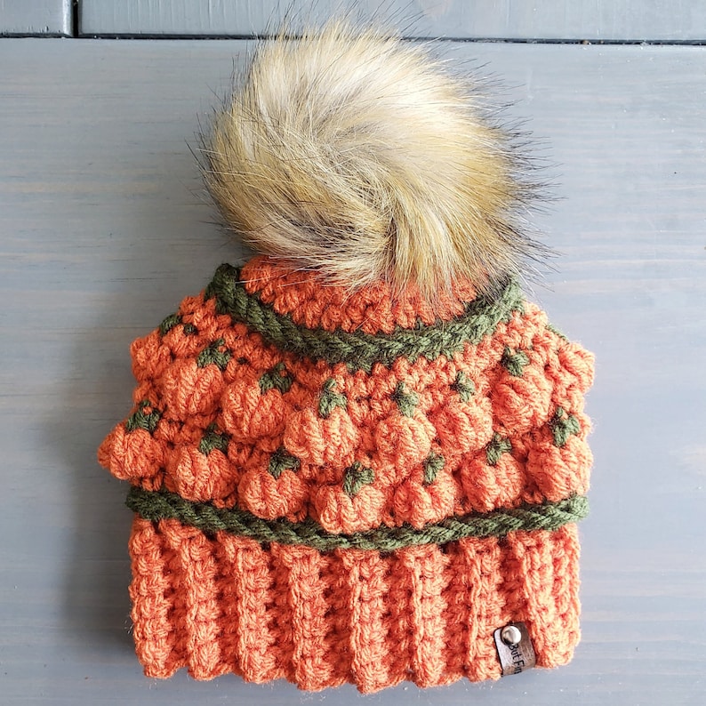 May include: Orange and green crocheted beanie with a fluffy brown pom pom on top. The beanie has a textured design that resembles pumpkins.