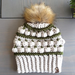 May include: A crocheted beanie hat with a faux fur pom pom. The hat is white with green stripes and a brown leather tag.