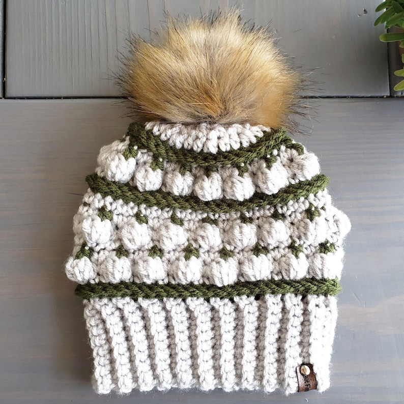 May include: A crocheted beanie hat with a faux fur pom pom. The hat is white with green stripes and a brown leather tag.