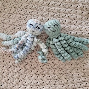 Puede incluir: Dos juguetes de pulpo hechos a mano con ganchillo. Uno es azul claro y blanco, el otro verde claro liso. Ambos tienen caras sonrientes y tent&aacute;culos rizados. Est&aacute;n colocados sobre una manta beige tejida.