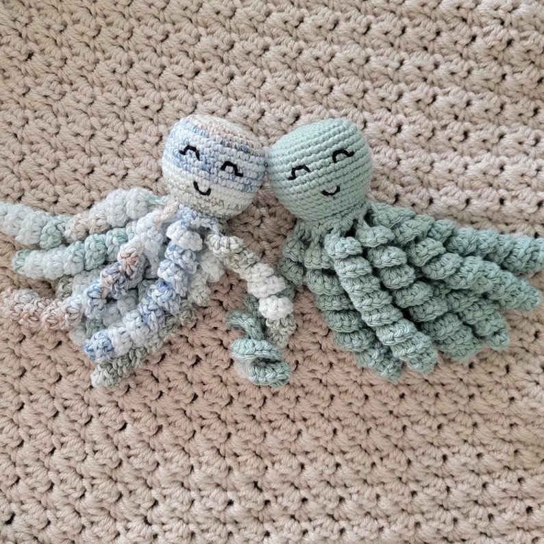 Puede incluir: Dos juguetes de pulpo hechos a mano con ganchillo. Uno es azul claro y blanco, el otro verde claro liso. Ambos tienen caras sonrientes y tent&aacute;culos rizados. Est&aacute;n colocados sobre una manta beige tejida.