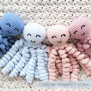 Op de afbeelding: Vier gehaakte octopus speelgoed in blauw, wit en roze, elk met gesloten ogen en een lachende mond. De speelgoedjes zijn gerangschikt op een getextureerd, gebroken witte ondergrond. De tekst "Crochet Pattern + Video" staat onderaan.