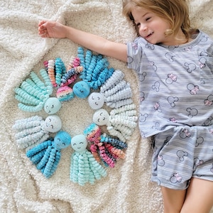 Puede incluir: Una colecci&oacute;n de coloridos juguetes de pulpo de ganchillo dispuestos en c&iacute;rculo sobre una manta blanca texturizada. Los pulpos presentan varios tonos de azul, rosa y verde. Cada pulpo tiene una cara sonriente. Los juguetes est&aacute;n hechos a mano.
