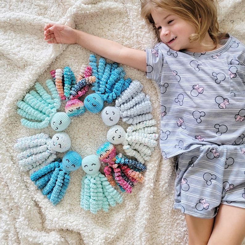 Puede incluir: Una colecci&oacute;n de coloridos juguetes de pulpo de ganchillo dispuestos en c&iacute;rculo sobre una manta blanca texturizada. Los pulpos presentan varios tonos de azul, rosa y verde. Cada pulpo tiene una cara sonriente. Los juguetes est&aacute;n hechos a mano.