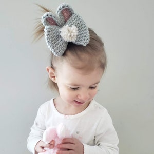K&ouml;nnte beinhalten: Ein grauer gestrickter Haarreifen mit Hasenohren und einem wei&szlig;en Bommel. Der Haarreifen wird von einem kleinen Kind mit blonden Haaren getragen.