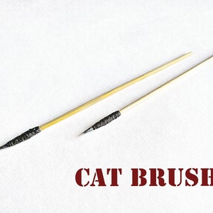 May include: Two wooden cat brushes with black bristles. The brushes are on a white surface. The text "CAT BRUSH" is printed in red below the brushes.