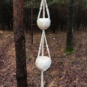 May include: A white macrame plant hanger with two white ceramic pots hanging from it. The hanger is suspended from a tree branch in a woodland setting.