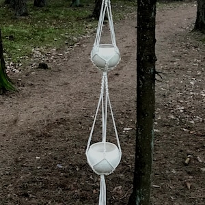 May include: A white macrame plant hanger with two white ceramic pots hanging from it. The hanger is suspended from a tree branch in a woodland setting.