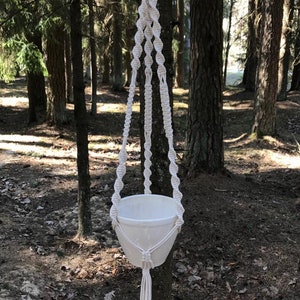 May include: A white macrame plant hanger with a white ceramic pot hanging from a wooden ring. The hanger is made of cotton cord and has a tassel at the bottom.
