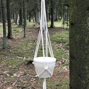 Peut inclure: Un porte-plante en macramé blanc avec un pot en céramique blanc suspendu à une branche d'arbre dans un environnement forestier.