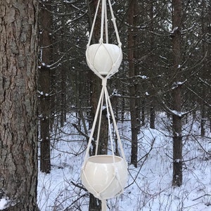 Op de afbeelding: Een dubbellaagse, crèmekleurige macramé plantenbak met twee witte keramische potten. De potten worden opgehangen door een geweven touwontwerp, met een houten ring aan de bovenkant om op te hangen. De achtergrond is een besneeuwd bos.