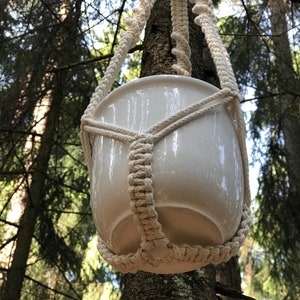 Op de afbeelding: Een witte keramische pot die hangt aan een macramé plantenhanger gemaakt van natuurlijk katoenen touw. De hanger is opgehangen aan een tak in een bosrijke omgeving.