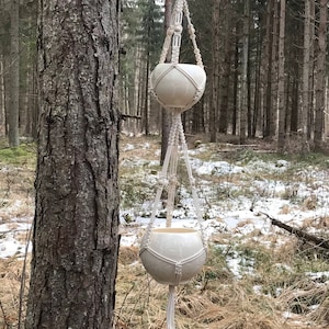 Op de afbeelding: Twee witte macramé plantenhangers met witte keramische potten die aan een tak hangen in een besneeuwd bos.