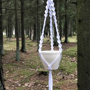 Könnte beinhalten: Ein weißer Makramee-Pflanzenhänger mit einem weißen Keramiktopf, der an einem Baum in einem Wald hängt.