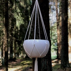 May include: White macrame plant hanger with a white ceramic pot hanging from a tree in a forest.