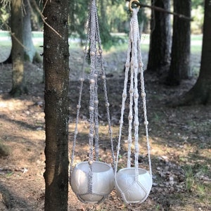 May include: Two macrame plant hangers with wooden beads hanging from a tree branch. The hangers are made of natural cotton cord and have a white ceramic pot at the bottom.