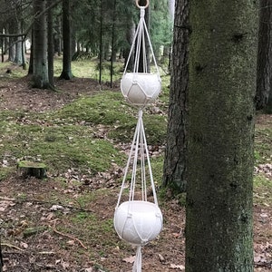 May include: A double-tiered, white macramé plant hanger with two round, cream-coloured ceramic pots. The hanger is suspended from a wooden ring and hangs in a forest setting, showcasing a natural, bohemian style. The pots have a textured design.