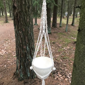 May include: White macrame plant hanger with a white ceramic pot hanging from a tree branch in a woodland setting.
