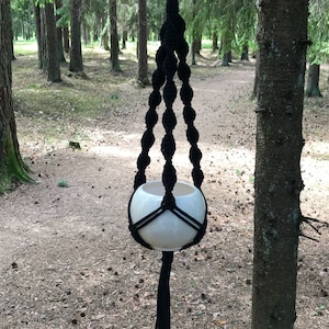 May include: Black macrame plant hanger with a white ceramic pot hanging from a tree branch in a woodland setting.