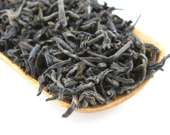 May include: Close-up of loose leaf tea on a wooden scoop. The tea leaves are dark green and brown, with a slightly curled appearance. The wooden scoop has a light brown color and is set against a white background. The tea appears to be a type of black tea.
