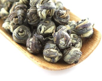 May include: A close-up of a wooden scoop filled with loose green tea pearls. The pearls are round and have a light green color with a white center. The tea pearls are arranged in a random pattern.