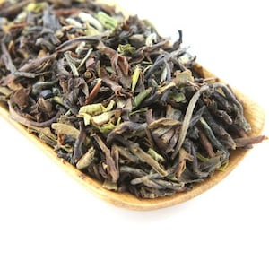 May include: Close-up of loose leaf tea in a wooden scoop. The tea leaves are a mix of dark brown, green, and tan, with a variety of textures. The wooden scoop is light brown and sits against a white background.