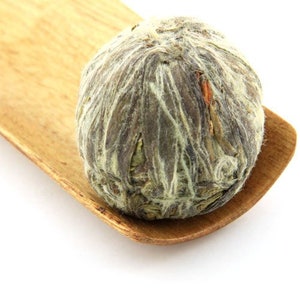 May include: A green tea flower bud, also known as a blooming tea, rests on a wooden spoon. The bud is tightly closed and has a light green color.