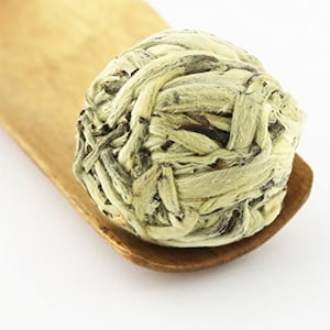 May include: A white tea ball, also known as a tea bloom, is a round ball of tea leaves that unfurls when steeped in hot water. The tea ball is made of white tea leaves and is resting on a wooden spoon.