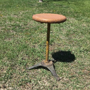 May include: A yellow and brown metal stool with a round seat and three legs. The stool is sitting on a grassy lawn.