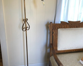May include: A gold-toned metal floor lamp with a curved arm and a decorative base. The lamp is in a room with a white wall and a wooden chair.