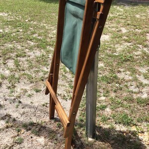 May include: A wooden folding chair with a green canvas seat. The chair is folded and leaning against a metal pole.