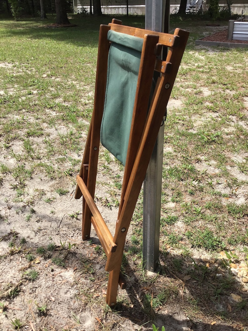 May include: A wooden folding chair with a green canvas seat. The chair is folded and leaning against a metal pole.