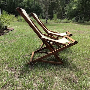 May include: Two wooden folding chairs with white canvas seats are sitting on a grassy lawn. The chairs are in a relaxed position, with the backs leaning back.