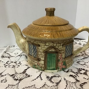 Puede incluir: Una tetera de cerámica con forma de cabaña con techo de paja. La tetera está decorada con pintura marrón, blanca y verde. La cabaña tiene una puerta verde y dos ventanas con cristales en forma de diamante.