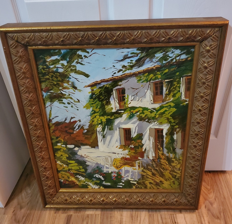 Puede incluir: Una pintura al &oacute;leo enmarcada que representa una casa blanca con enredaderas verdes que crecen por el lado. La casa tiene ventanas y una puerta, y hay &aacute;rboles y flores en primer plano. La pintura est&aacute; en un marco dorado.