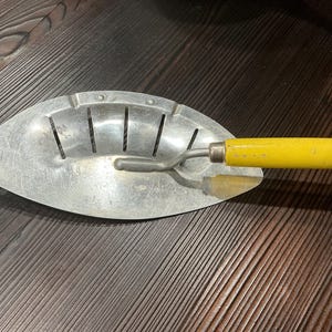 May include: A vintage metal kitchen strainer with an oval-shaped, perforated bowl and a bright yellow wooden handle. The silver-toned metal surface features a series of vertical slots for draining liquids and a small circular hole at the pointed end.